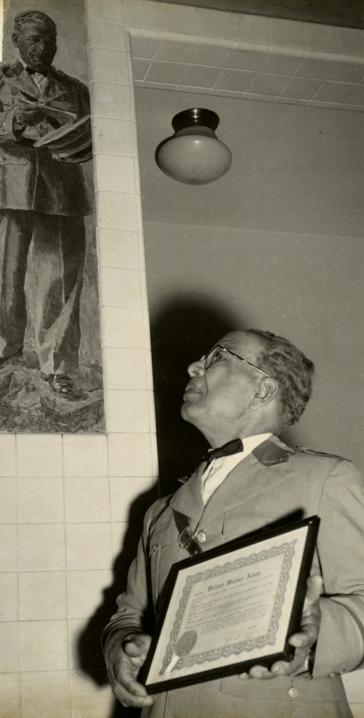 A black and white picture of Wallace Alsop standing in front of a depiction of himself in the George Washington Hall mural. He wears glasses, has short hair, and his dressed in a suit complete with a bow tie. In his hands he has a Meritorious Service Certificate that was presented to him at a ceremony held in honor of his retirement in 1956.