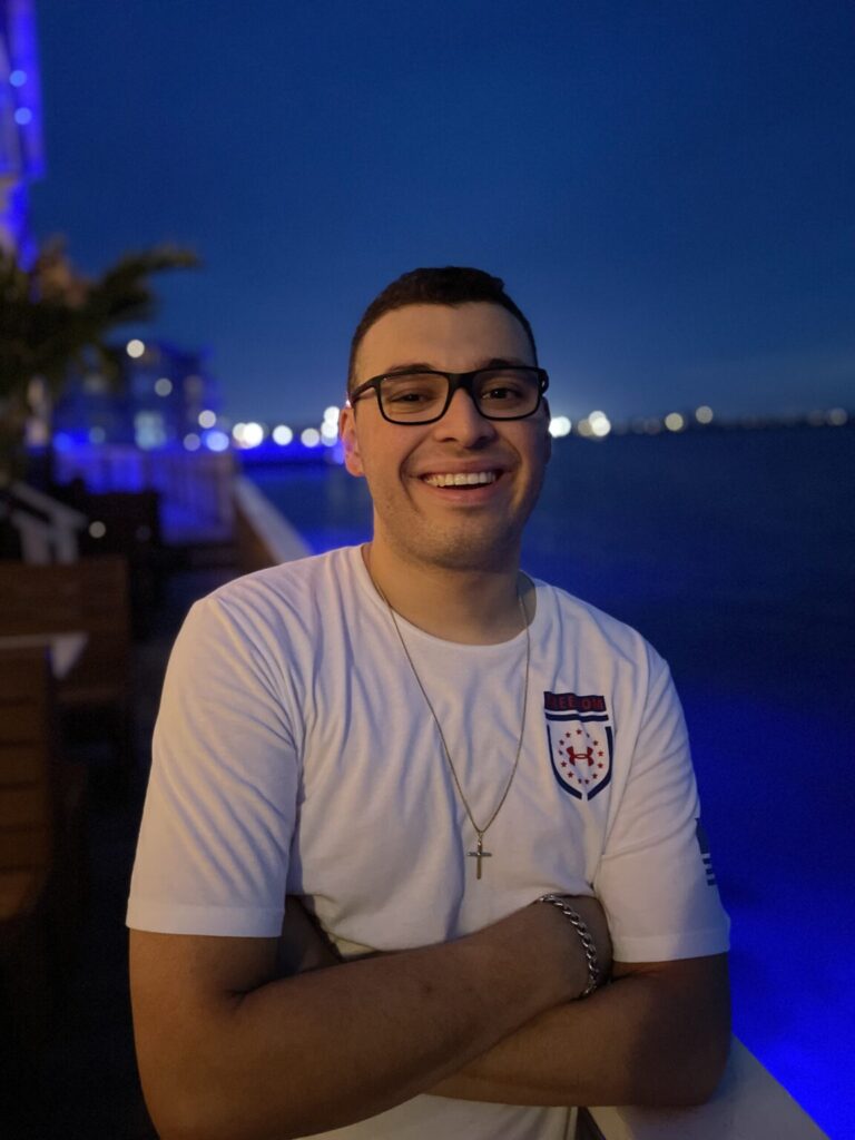 A picture of Bryan Rivas. He is Caucasian and has his arms crossed in front of him. He has short black hair and black glasses. His shirt is white with a logo on the top right and he has a cross around his neck. A silver chain is around his right wrist. He appears to be by a body of water at night.