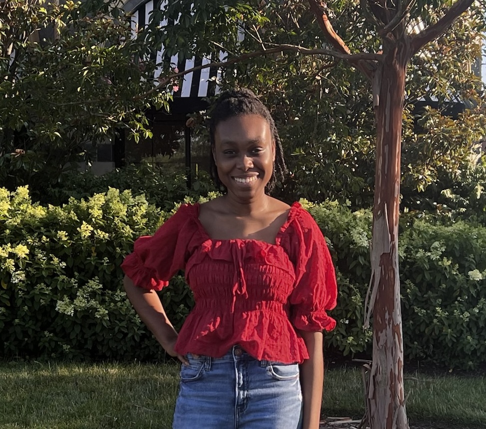 A picture of Abby Delapenha. She is African American. The photo was taken outdoors and she is by a tree. She wears a red top and jeans. Her hair is pulled back out of her face and she is smiling widely at the camera. 