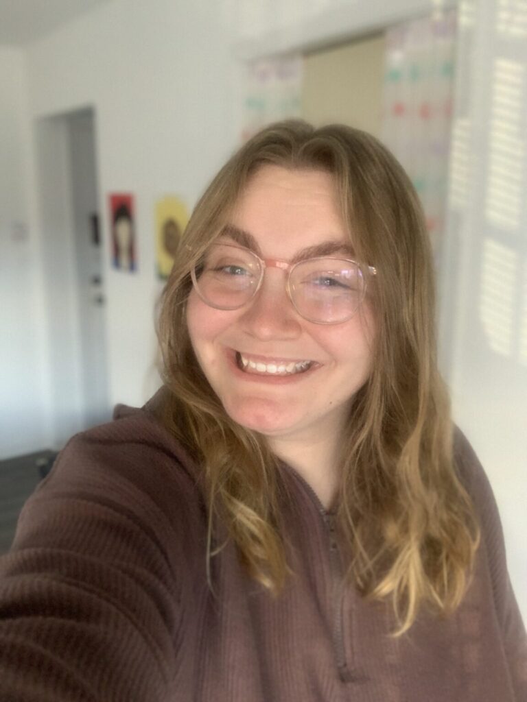 A picture of Taylor Coleman. She is Caucasian, has medium length dirty-blonde hair parted in the center, and pale pink glasses. She wears a brown hoodie. She is giving a toothy grin to the camera. The background behind her is blurred.
