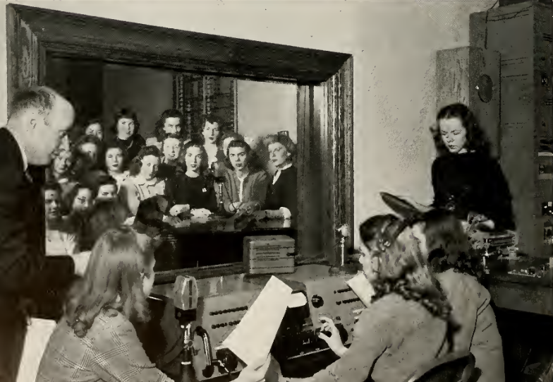 A black and white image of the Station WMWC. It shows three women recording into microphones, a man to their left. Another woman is behind a desk to the right. On the wall is a picture of a group of people.