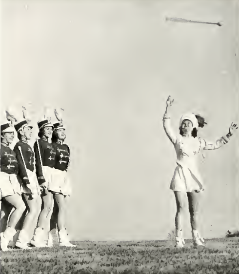 A black and white photo from 1952 shows four marching women to the left holding majorette batons. To the right is a marcher in white looking ready to catch a baton that is out of range of the photograph.